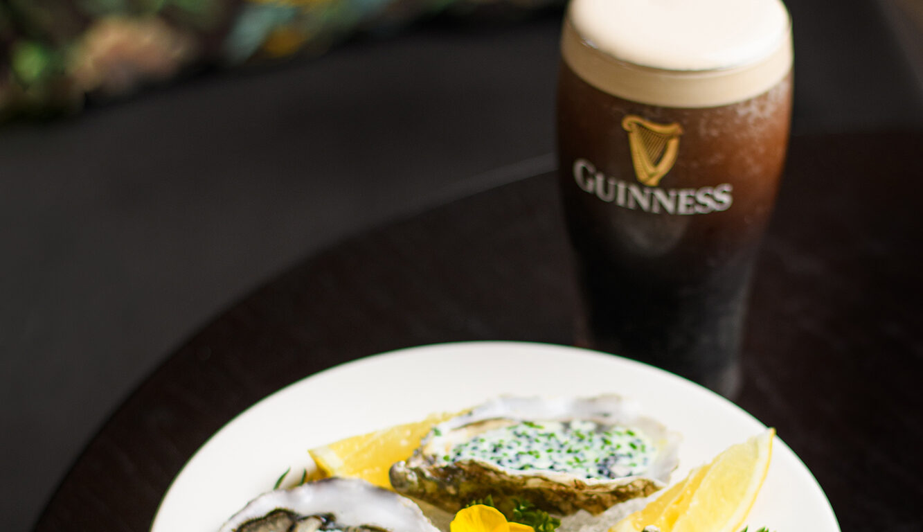 A plate of three oysters garnished with green herbs and lemon wedges, served alongside a pint of Guinness stout on a dark table.
