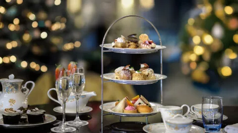 Three-tiered tray with assorted pastries and sandwiches on a table, alongside teacups, a teapot, desserts, and glasses of champagne, with blurred festive lights in the background.