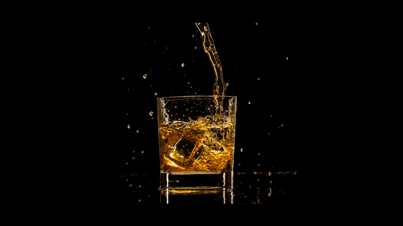 A glass filled with amber liquid and ice cubes, with more liquid being poured in, against a black background with visible splashes and droplets.