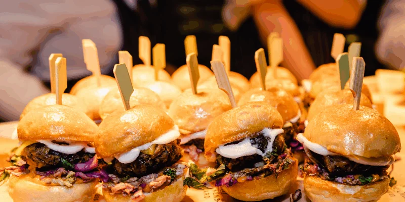 A row of mini sliders with patties, coleslaw, and sauce, each held together with a small wooden skewer, arranged on a tray.