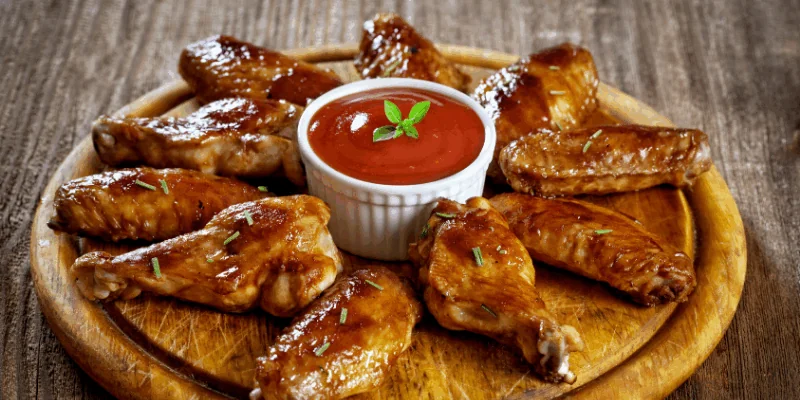 A wooden platter with glazed chicken wings arranged in a circle around a small dish of red dipping sauce, garnished with a sprig of green herb.