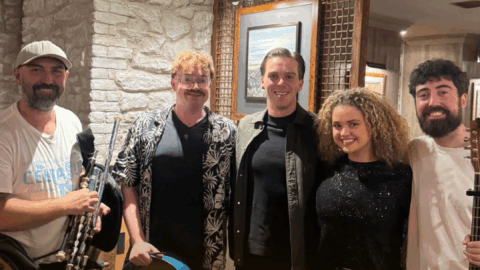 Five people stand indoors, two holding musical instruments: one with Uilleann pipes and another with a guitar. They are smiling and appear to be in a casual, warmly lit setting.