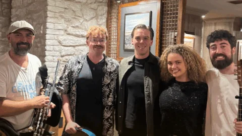 Five people stand indoors, two holding musical instruments: one with Uilleann pipes and another with a guitar. They are smiling and appear to be in a casual, warmly lit setting.