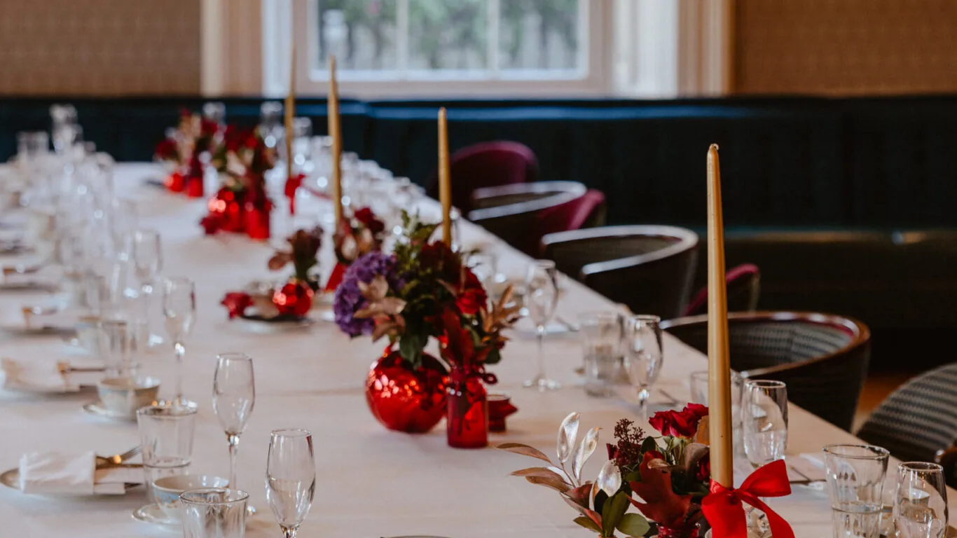 A long dining table set with white tablecloth, red and gold centerpieces, tall candles, glassware, and teacups in a restaurant with large windows and framed art on the walls.