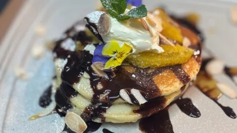 A stack of pancakes topped with cream, orange slices, edible flowers, mint leaves, sliced almonds, powdered sugar, and drizzled with chocolate sauce on a plate.