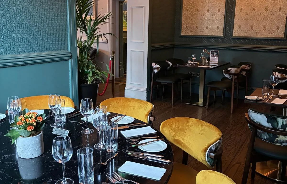 Elegant restaurant dining room with a round table set for six, yellow velvet chairs, glassware, and cutlery; wall art and chandelier in the background.