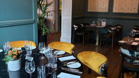 Elegant restaurant dining room with a round table set for six, yellow velvet chairs, glassware, and cutlery; wall art and chandelier in the background.