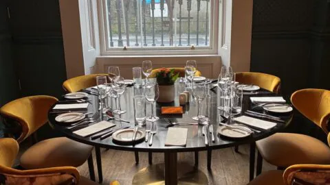 A round dining table set for six with plates, glasses, and cutlery, surrounded by yellow chairs, placed near a window with a street view outside.