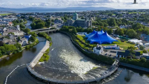Scenic aerial view of Galway city with river, lush greenery, iconic blue tent, and historic cathedral under a bright blue sky.