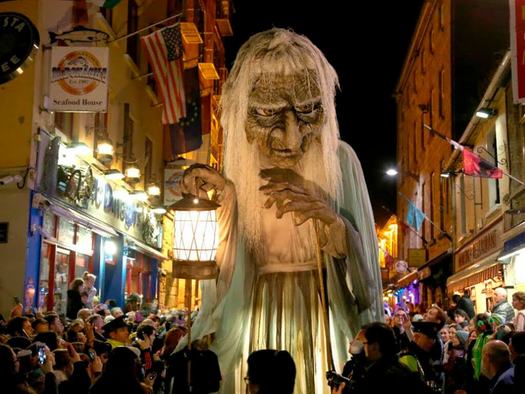 Bustling street festival with giant folklore figure in vibrant evening, surrounded by joyful crowd and lively shopfronts.