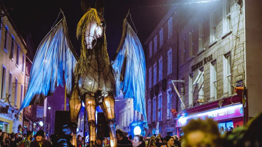 Night parade with glowing Pegasus float, vibrant crowd, and charming stone buildings in a festive atmosphere.